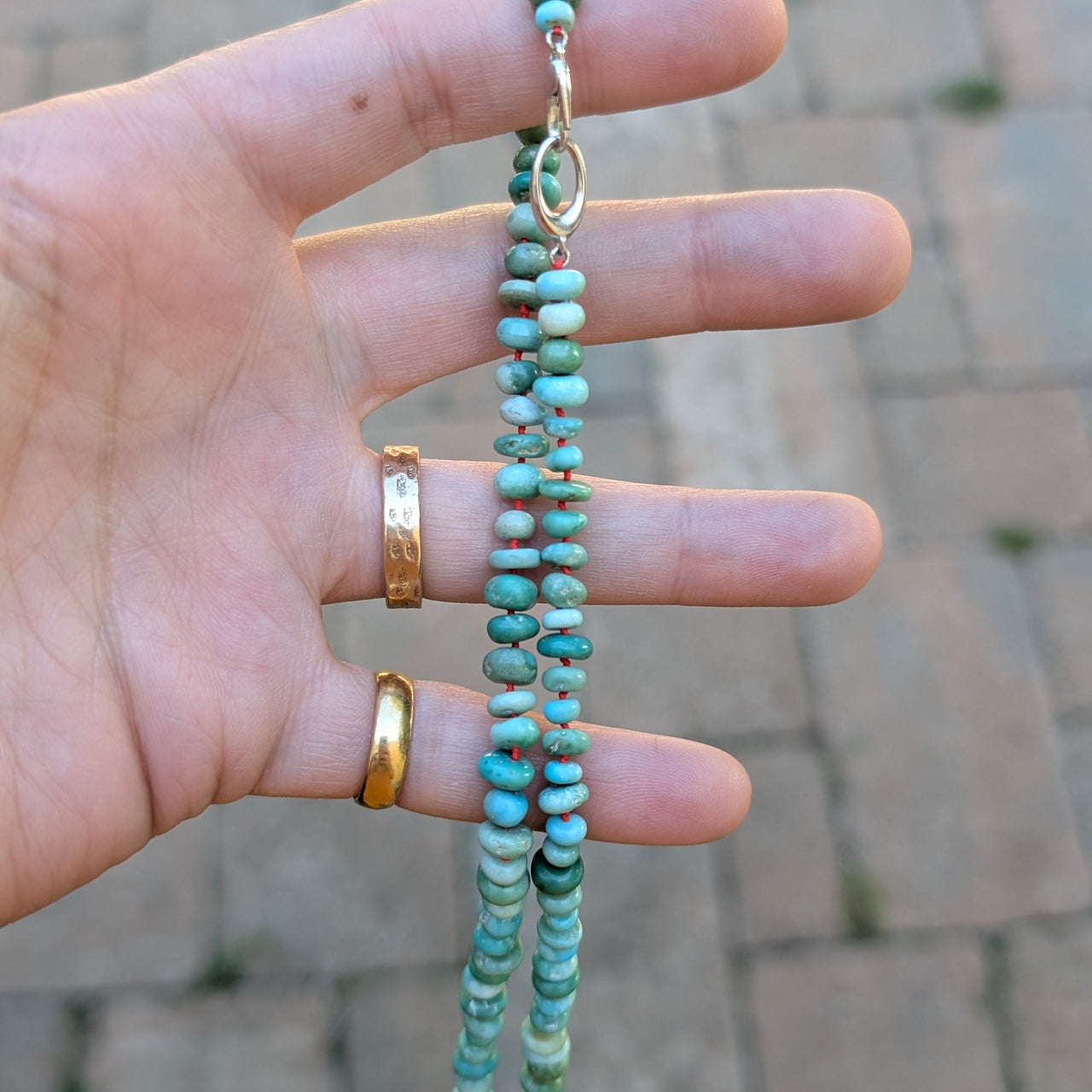 Turquoise beaded necklace held by a hand with gold rings, blurred brick pavement background